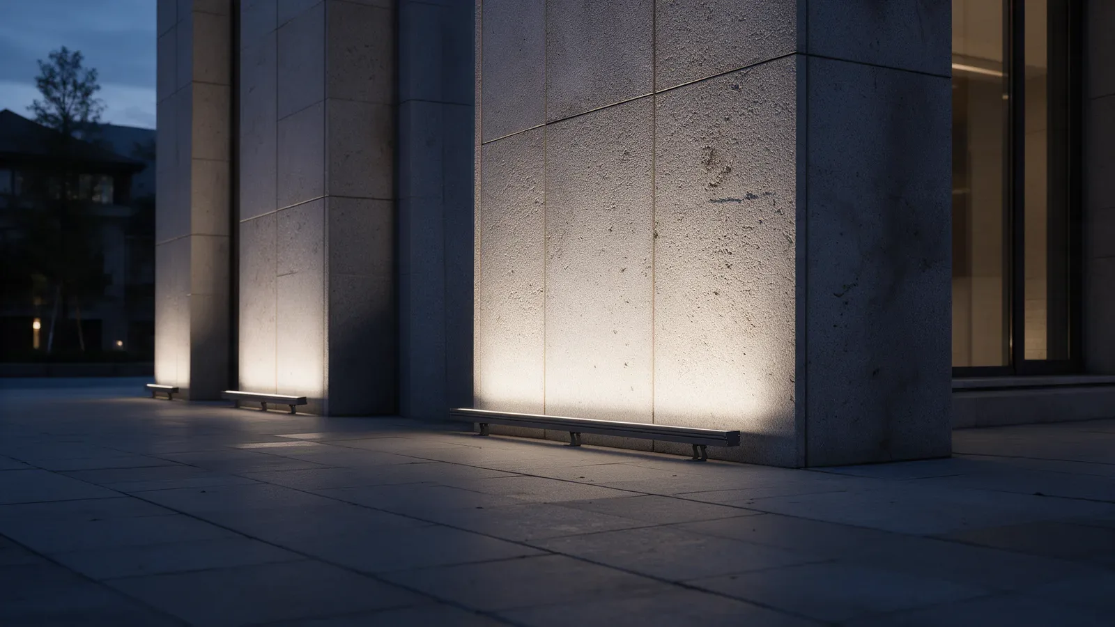 Fachada de un edificio público de piedra texturizada al atardecer, con barras de luz discretas al pie del muro.