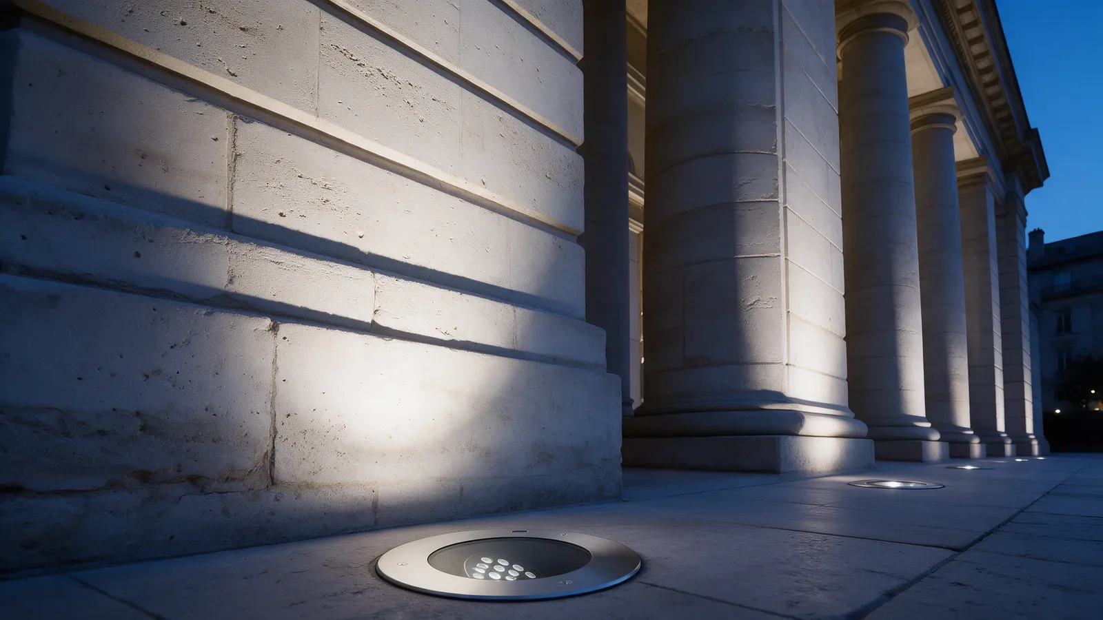 Fachada de un edificio público de piedra caliza al anochecer, iluminada por proyectores empotrados al pie de las columnas.